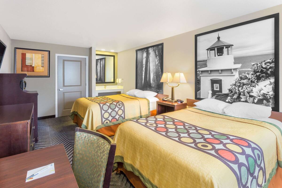 A hotel room with two beds, a desk, a chair, a desk lamp, a nightstand, and a large framed picture on the wall.