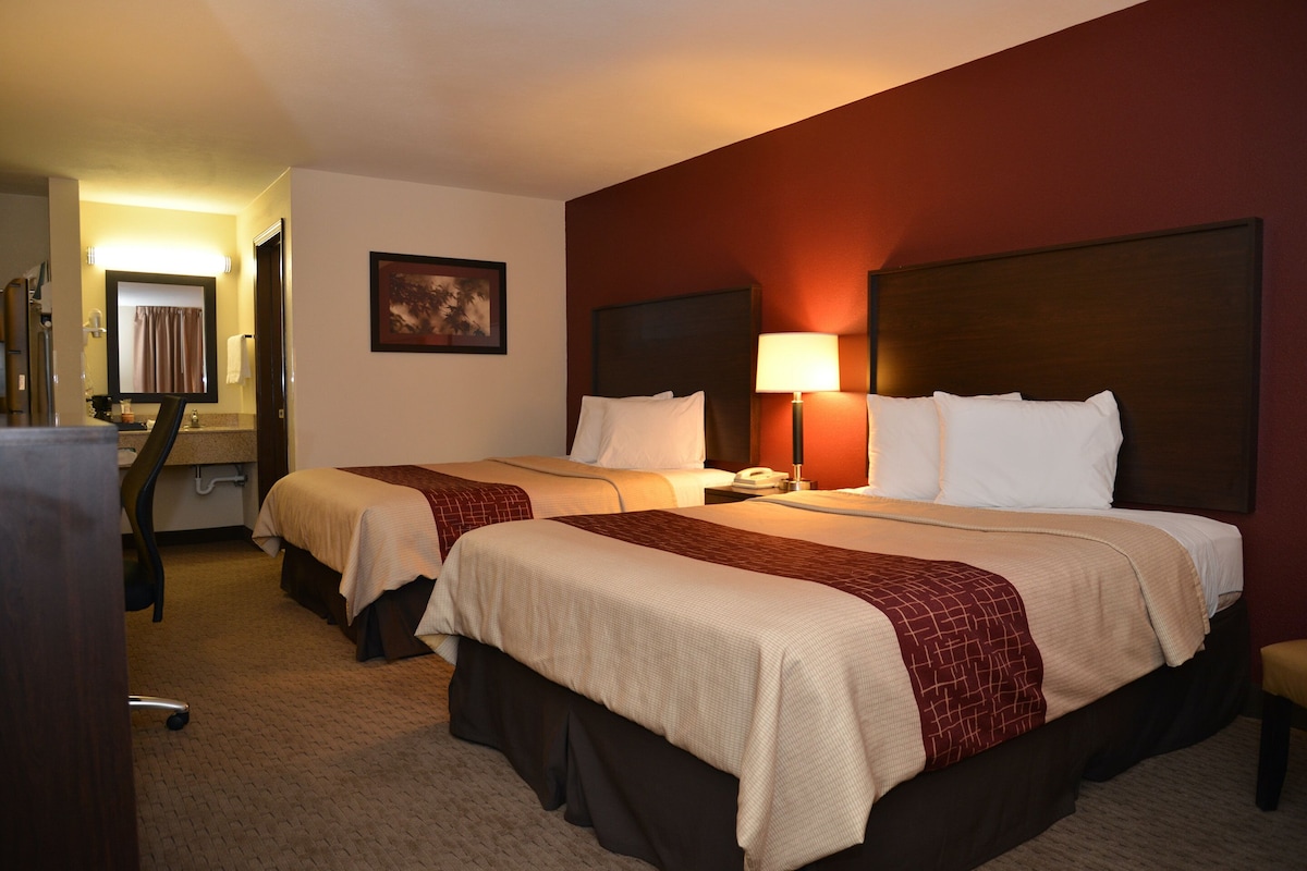 A hotel room with two beds, a desk, a chair, a lamp, and a painting on the wall.