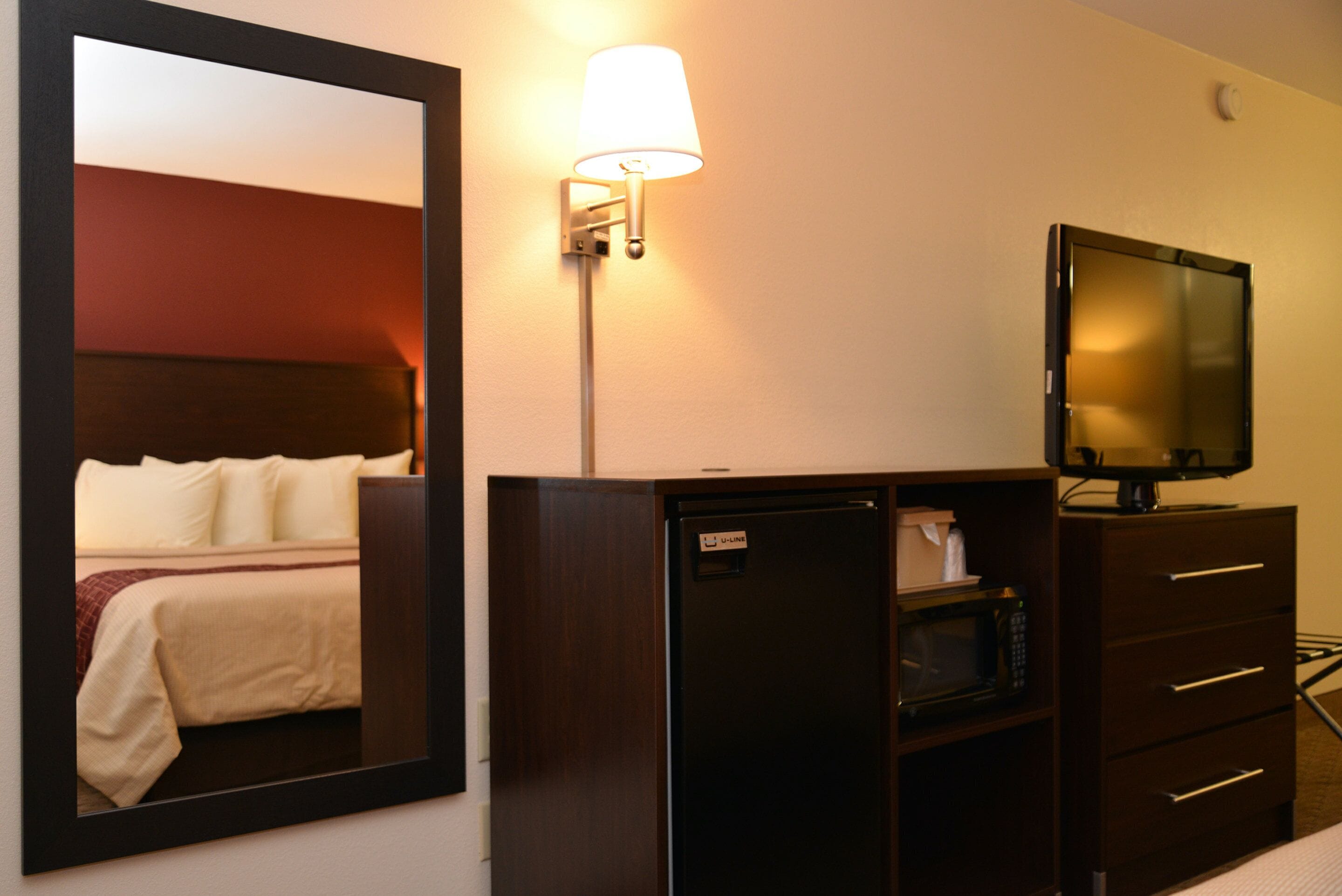 A hotel room with a bed, a television on a cabinet, a refrigerator, and a wall-mounted lamp.