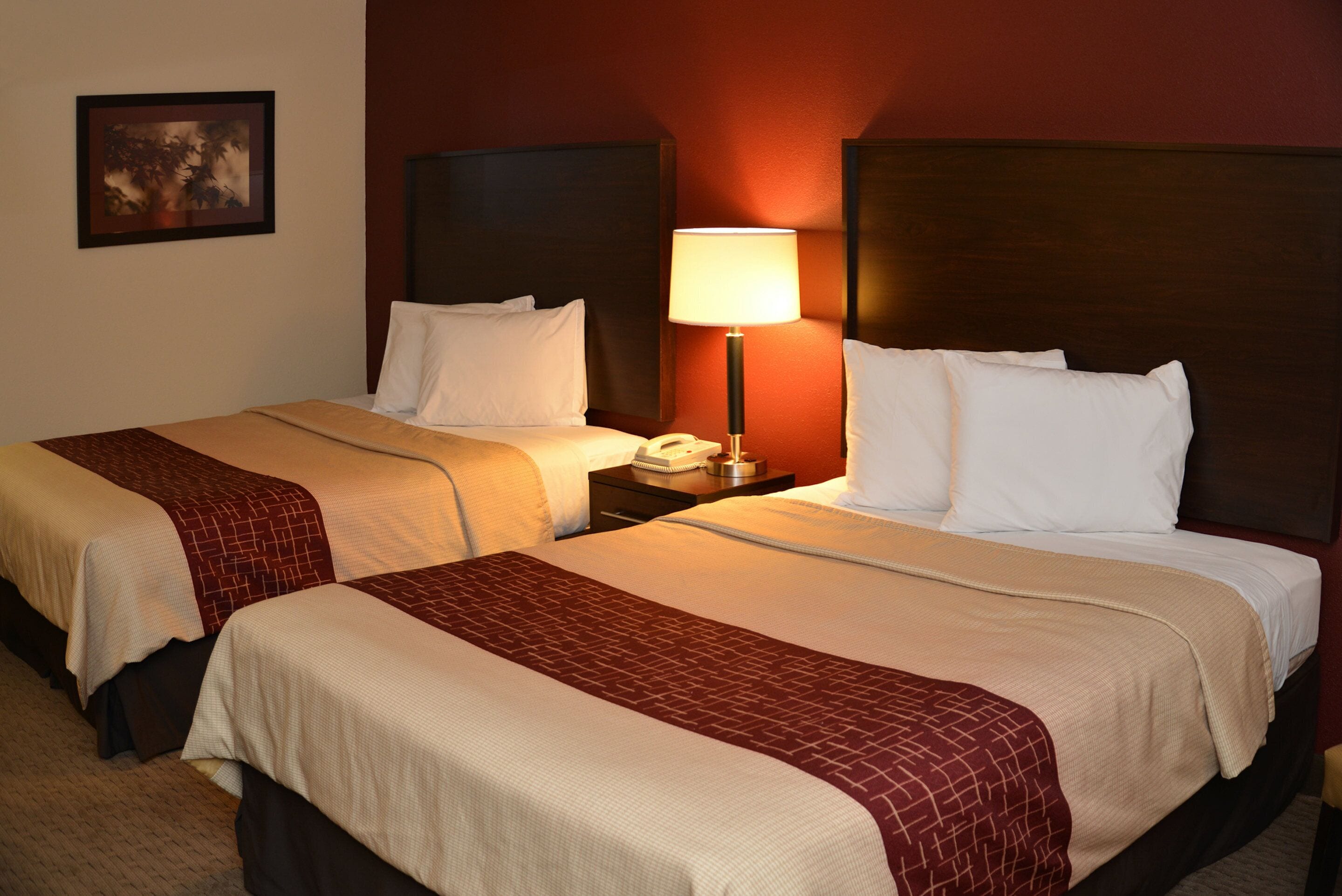 A hotel room with two beds, each with a headboard, a bedside table with a lamp, and a framed picture on the wall.