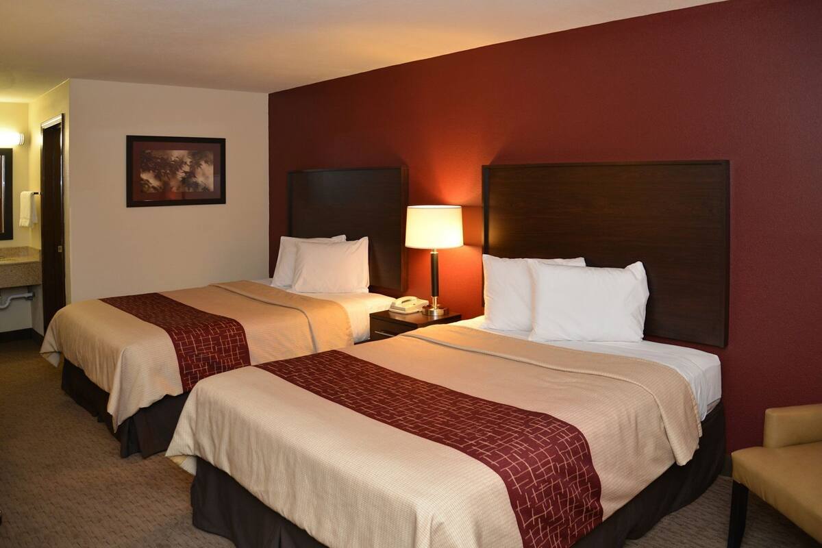 A hotel room with two beds, a nightstand with a lamp, a painting on the wall, and a carpeted floor.