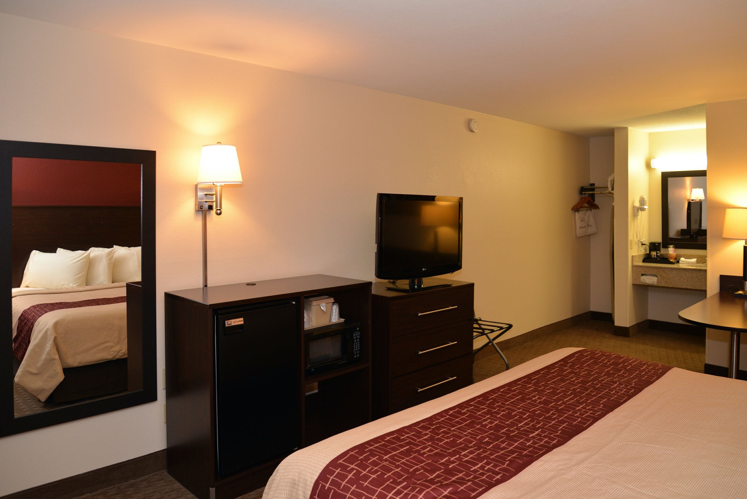 Habitación de hotel con cama, televisión, escritorio, espejo y baño visible a través de una puerta abierta.