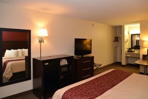 Habitación de hotel con cama, televisión, escritorio, espejo y baño visible a través de una puerta abierta.