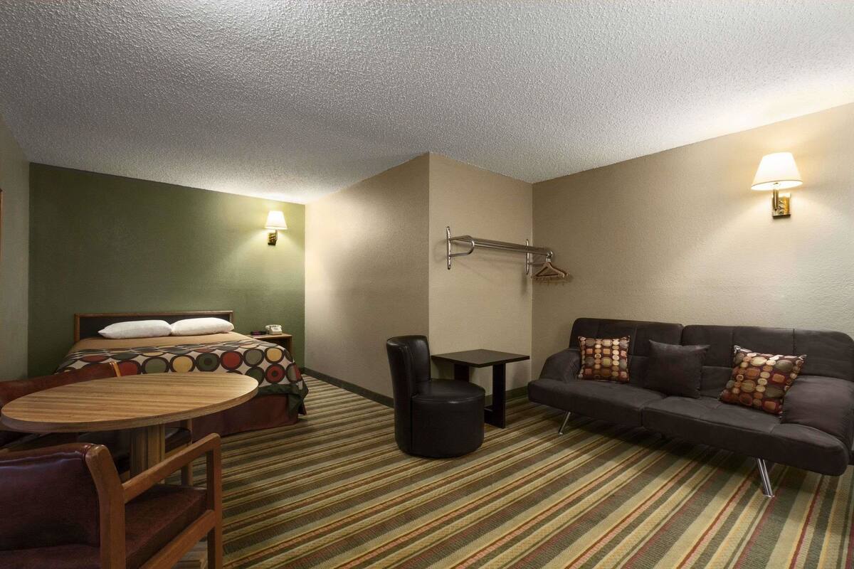 Habitación de hotel con una cama, un sofá, una mesita y una silla. Hay una lámpara en la pared y un perchero.