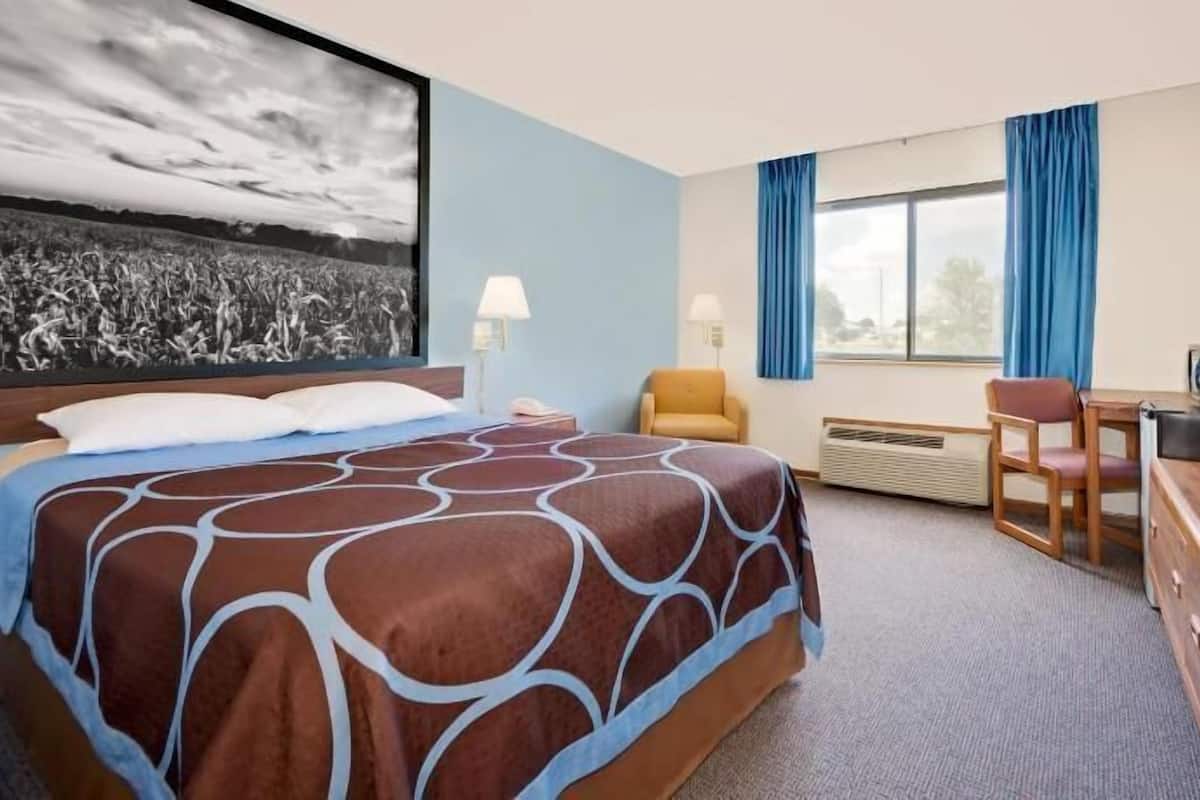 Una habitación de hotel con una cama, un escritorio con televisor, una silla y un ventanal amplio.