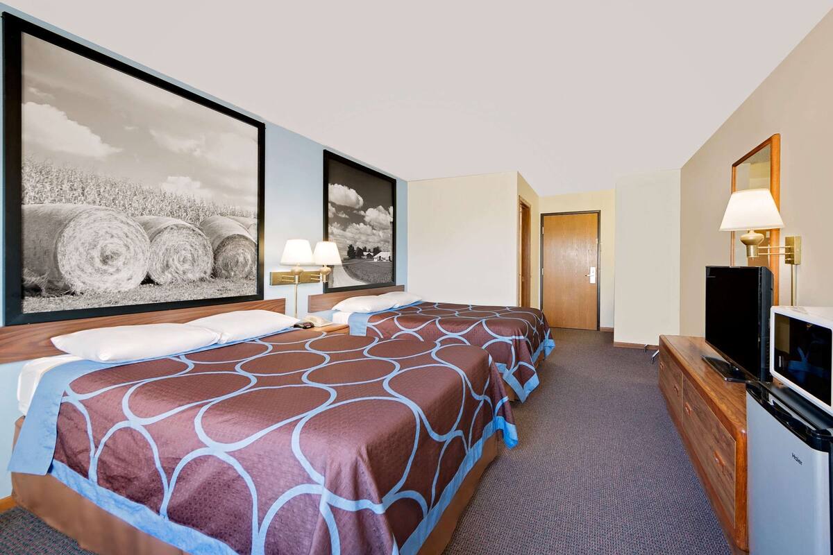 A hotel room with two beds, a television, a desk with a lamp, and large framed pictures on the wall.