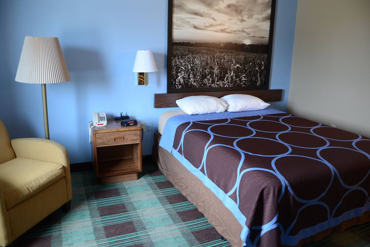 A hotel room with a bed, a chair, a nightstand with a lamp, and a wall-mounted telephone.