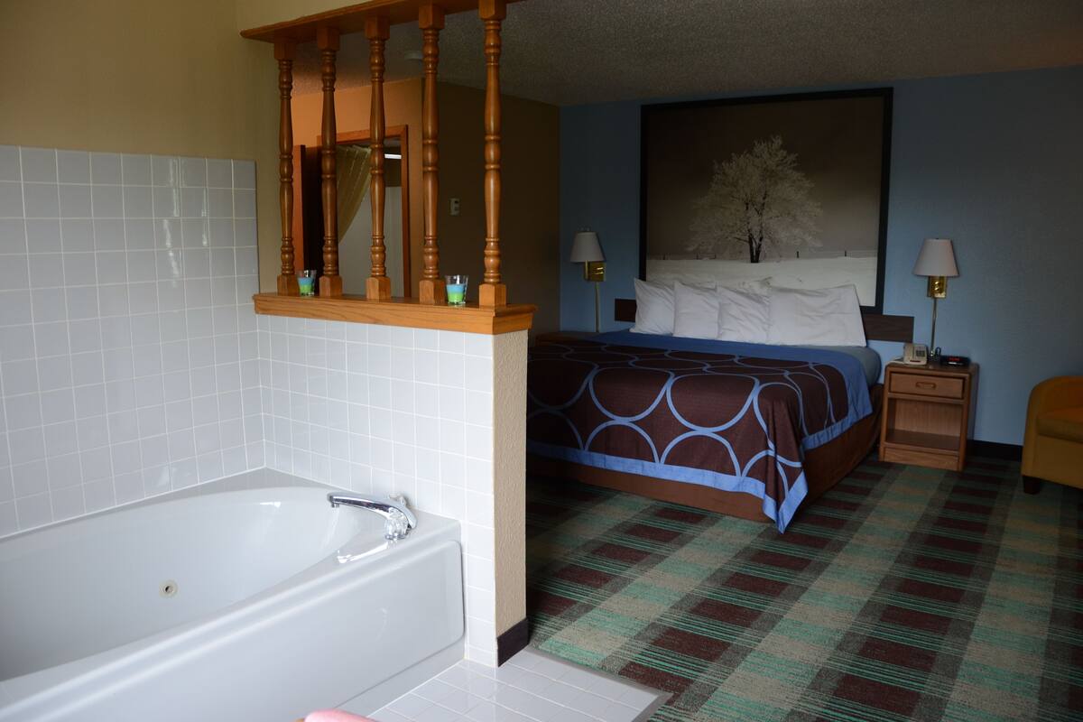 A hotel room with a large white bathtub, a bed with a blue and brown patterned bedspread, and a nightstand with two lamps.