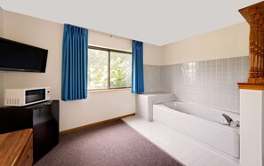 A bathroom with a white bathtub, a flat-screen TV mounted above a white microwave, and blue curtains on a window.