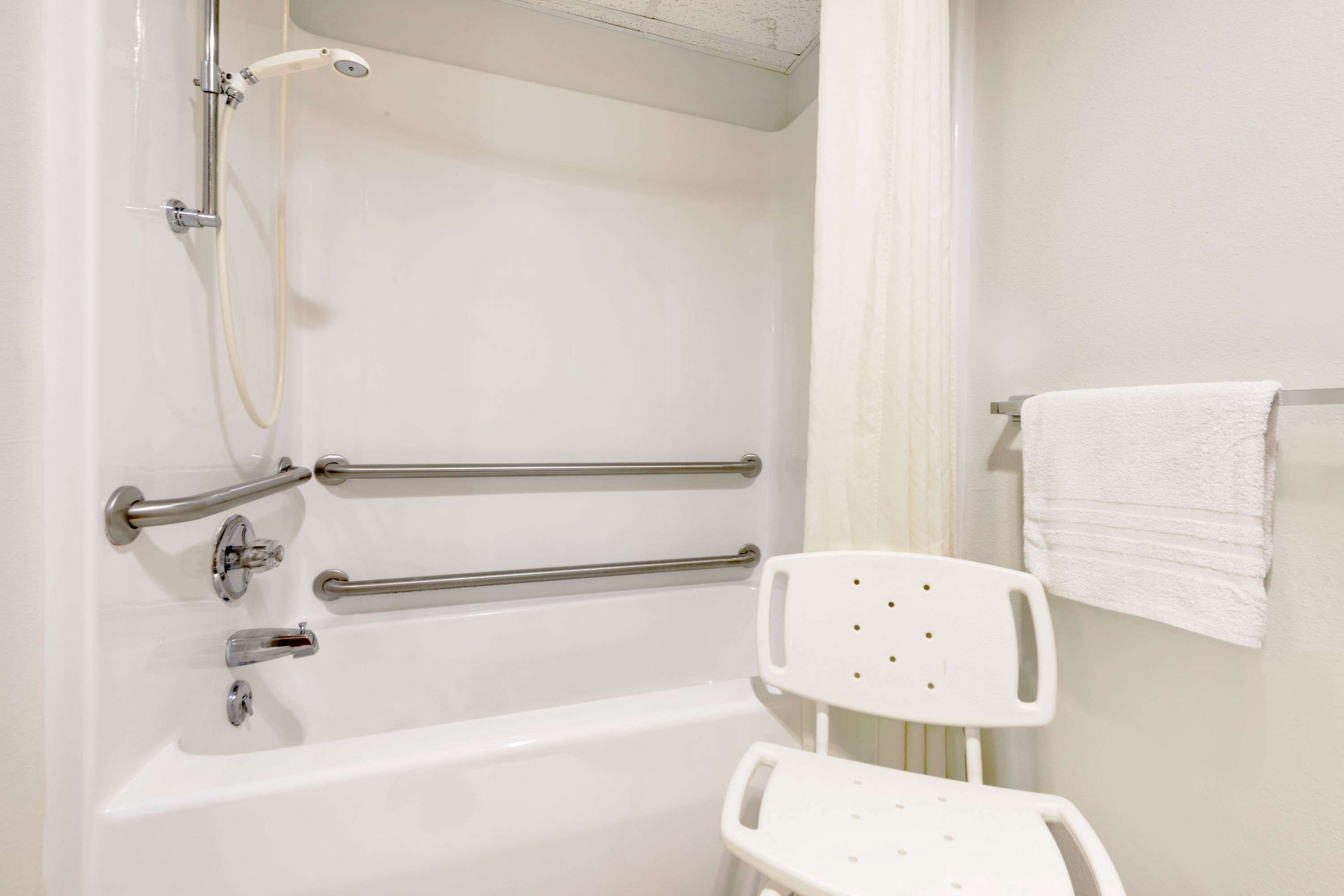 Une salle de bain équipée d’une baignoire, de barres d’appui, d’une chaise blanche et d’un porte-serviettes.