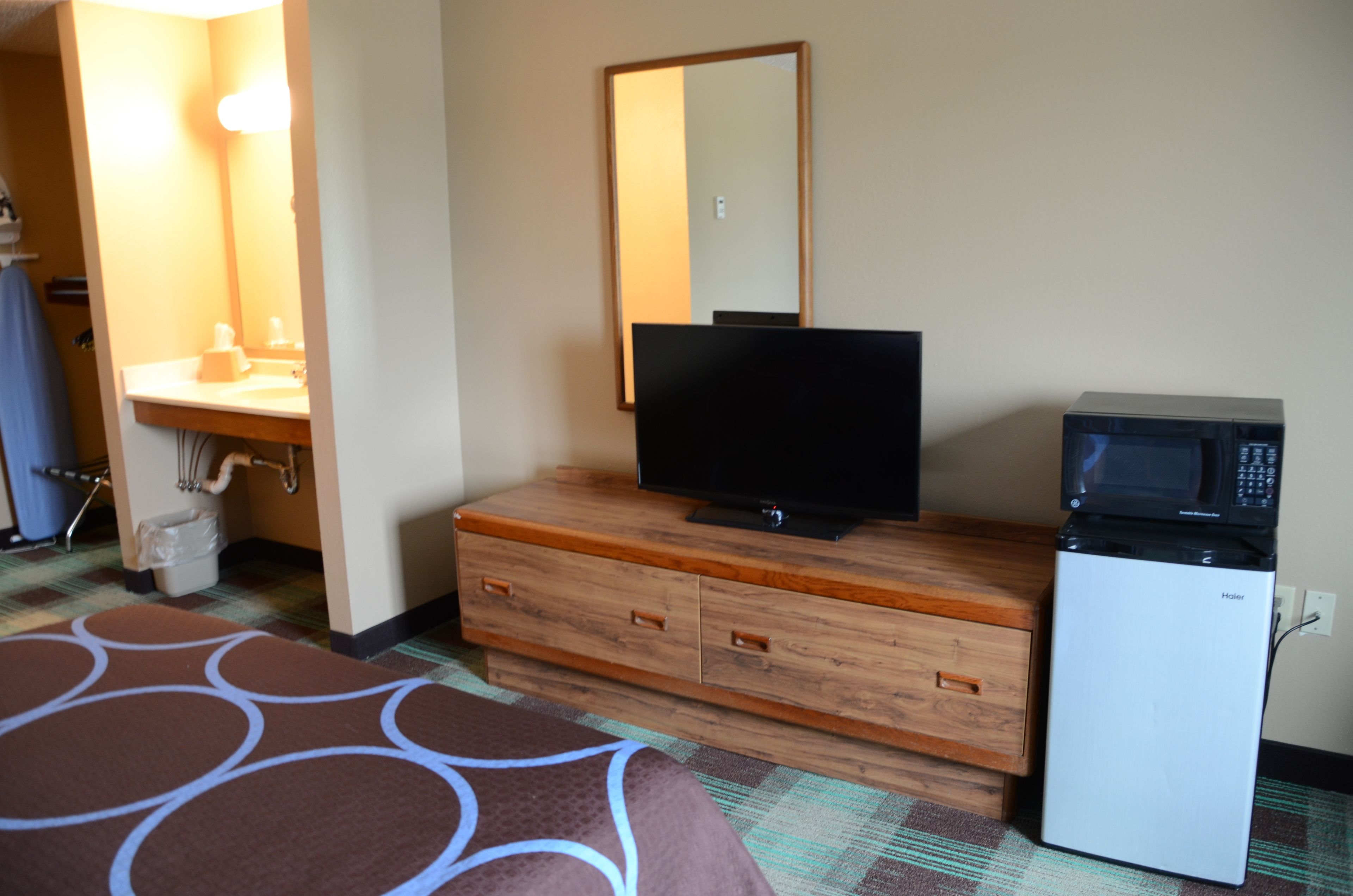 A hotel room with a wooden dresser, a television, a microwave, and a bathroom with a sink and mirror.