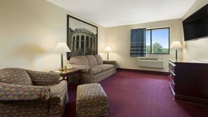A hotel room with a sofa, armchair, ottoman, a flat-screen TV, a desk with a lamp, a framed picture on the wall, and a window with curtains.