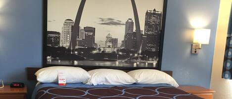A hotel room with a bed, a nightstand, a clock, a wall-mounted lamp, and a cityscape poster.
