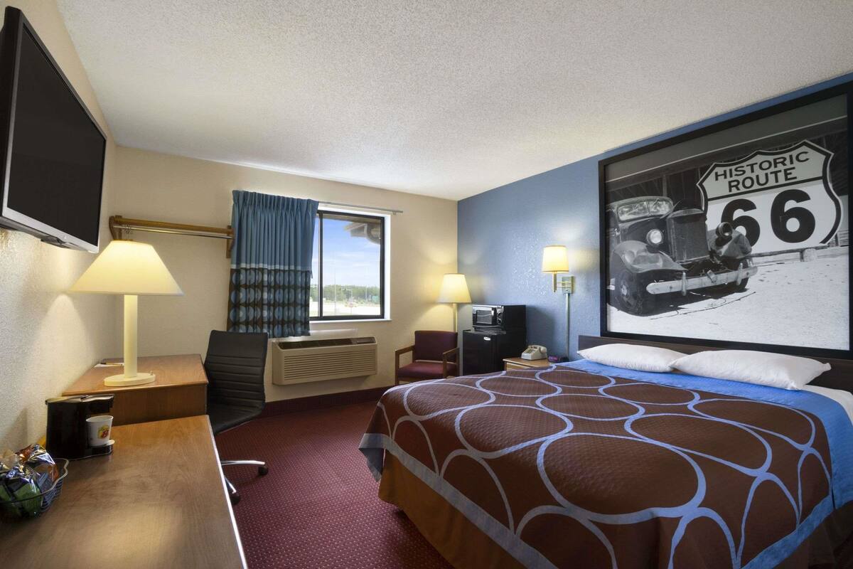 A hotel room with a bed, desk, and chair. There is a window with curtains, a television, and a wall-mounted lamp.