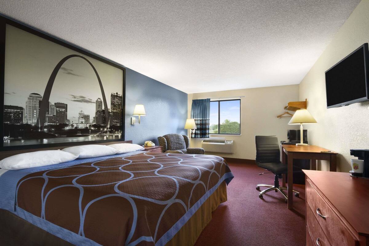 A hotel room with a large bed, a desk with a computer, a chair, a TV, and a window with a view of buildings.