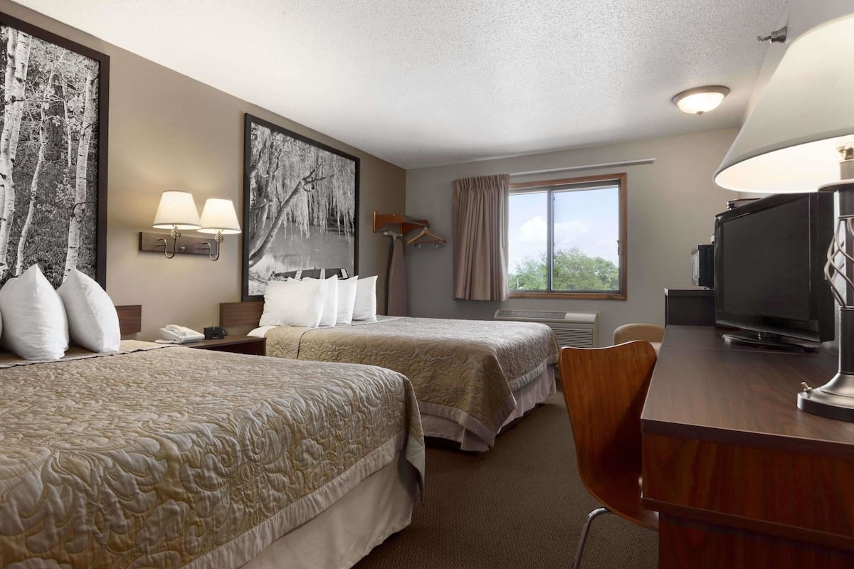 A hotel room with two beds, a desk with a TV, a lamp, and a large window.
