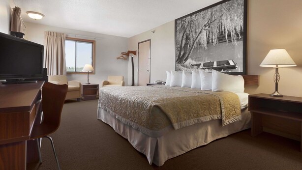 Habitación de hotel con cama, escritorio, silla, televisor, ventana con cortinas, lámpara y un cuadro grande enmarcado en la pared.