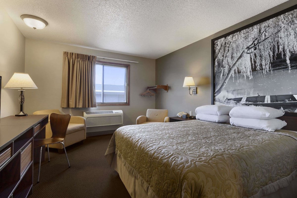 A hotel room with a large bed, a desk with a chair, a window with curtains, a wall-mounted lamp, and a large framed picture.