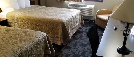A hotel room with two beds, a desk, a chair, a TV, a window with curtains, and a wall-mounted heater.