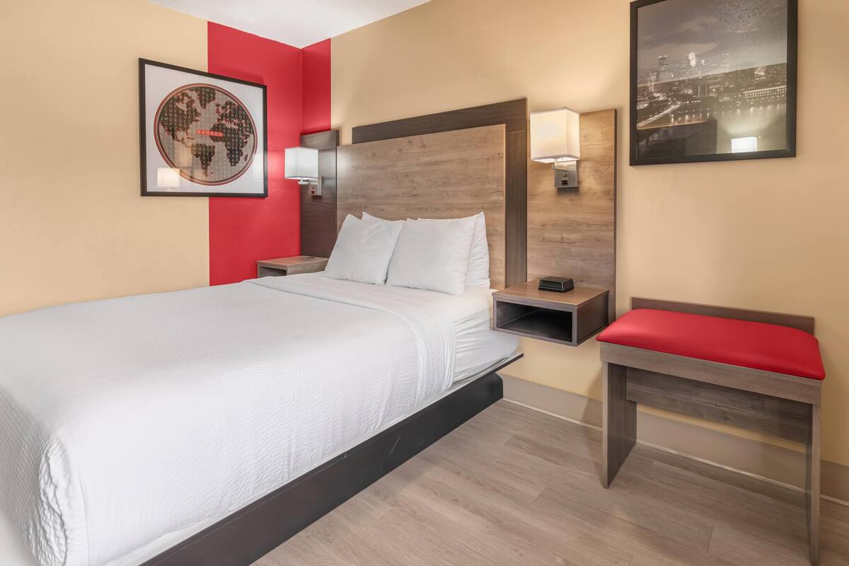 A hotel room with a bed, a nightstand, a red bench, a wall-mounted lamp, and a framed picture of a cityscape.
