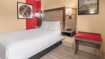 A hotel room with a bed, a nightstand, a red bench, a wall-mounted lamp, and a framed picture of a cityscape.