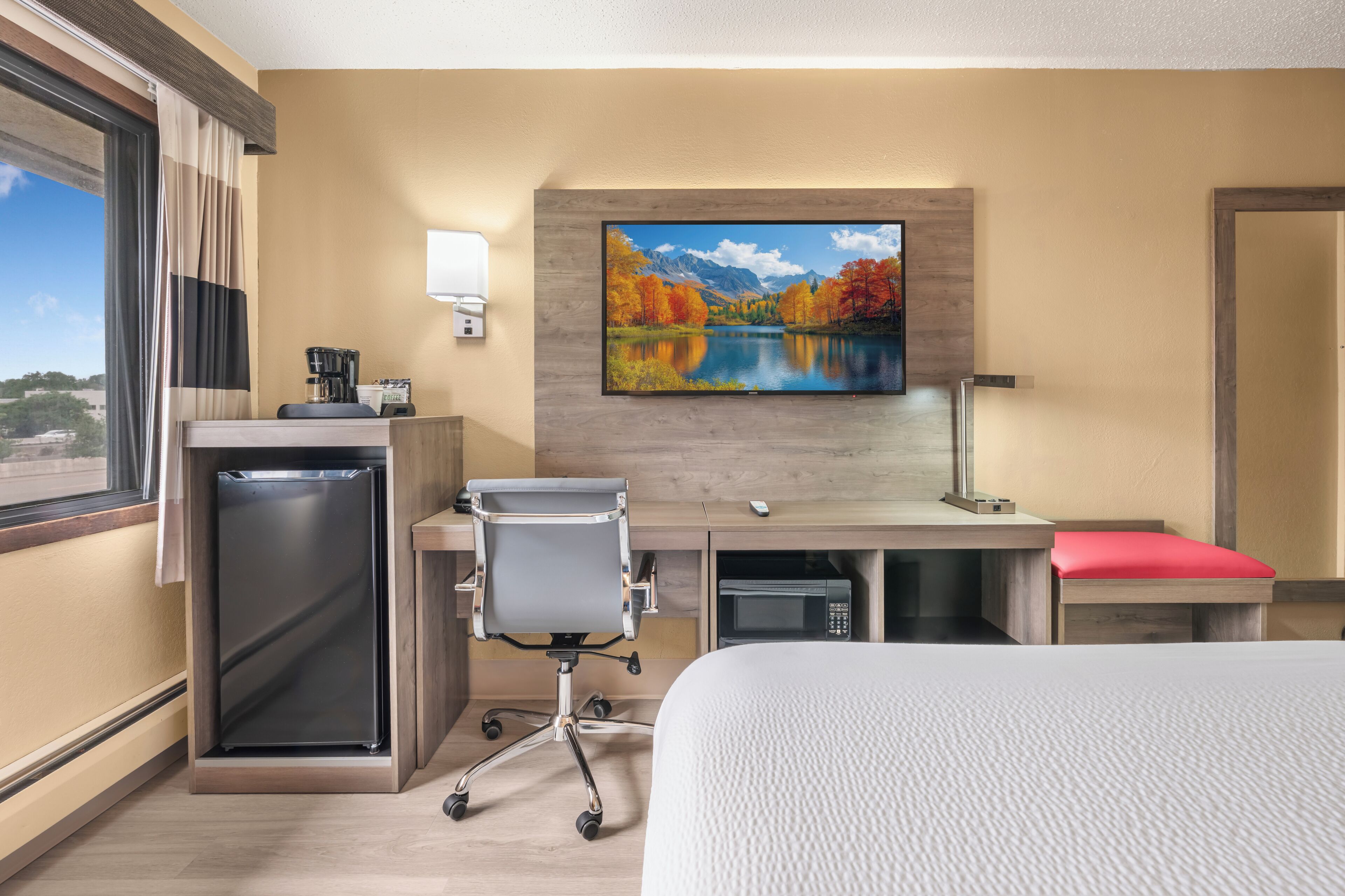 A hotel room with a bed, desk, chair, microwave, and a painting of a lake and mountains.