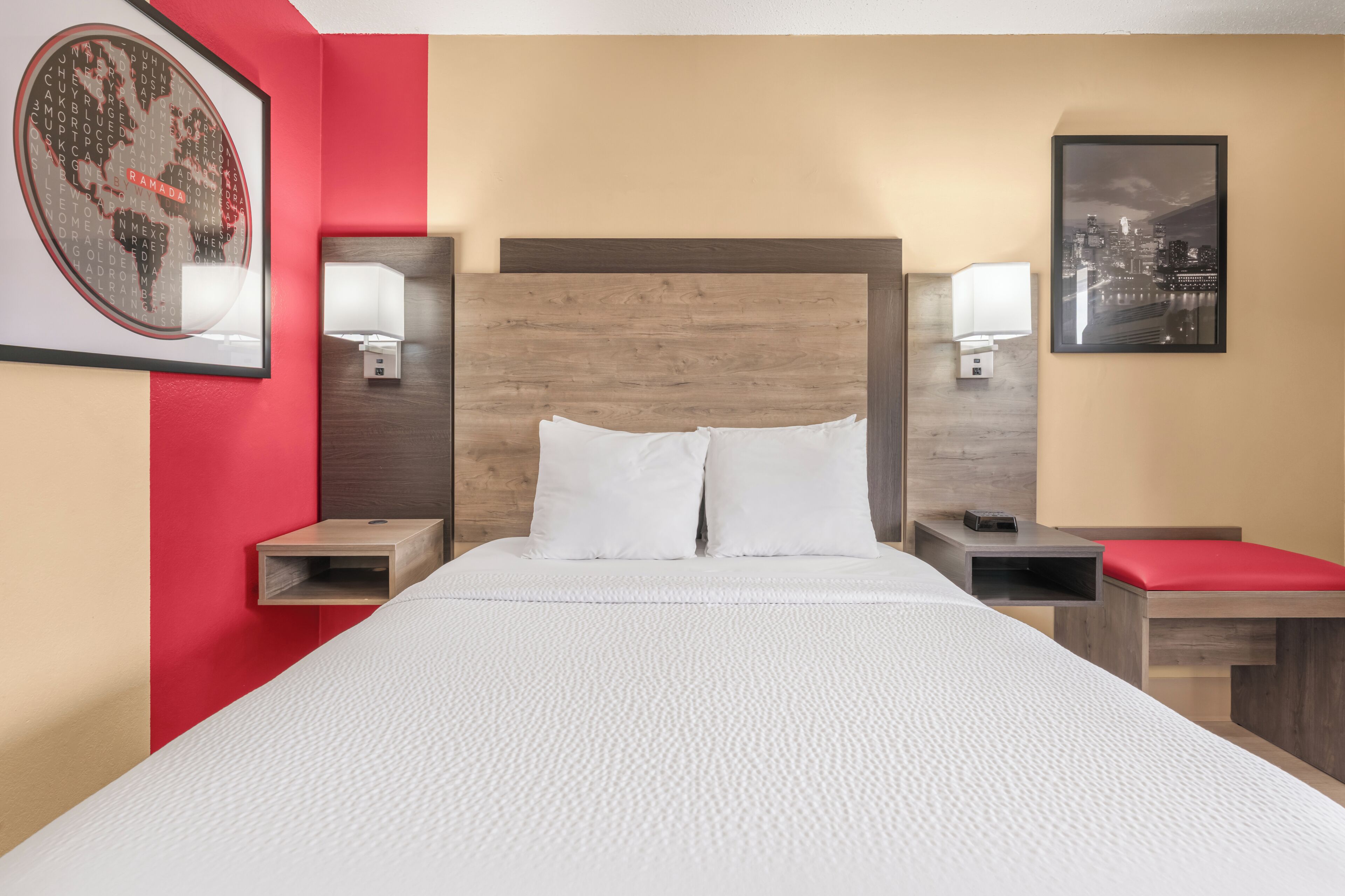 Une chambre d’hôtel avec un grand lit, deux tables de chevet, un banc rouge, une tête de lit en bois et deux lampes fixées au mur.