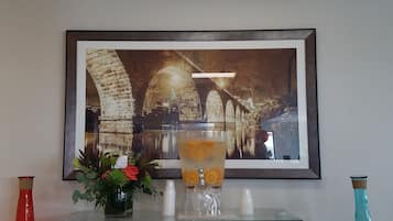 Una obra de arte enmarco que muestra un puente arqueado, una mesa de vidrio con una bebida y dos jarrones sobre una consola.