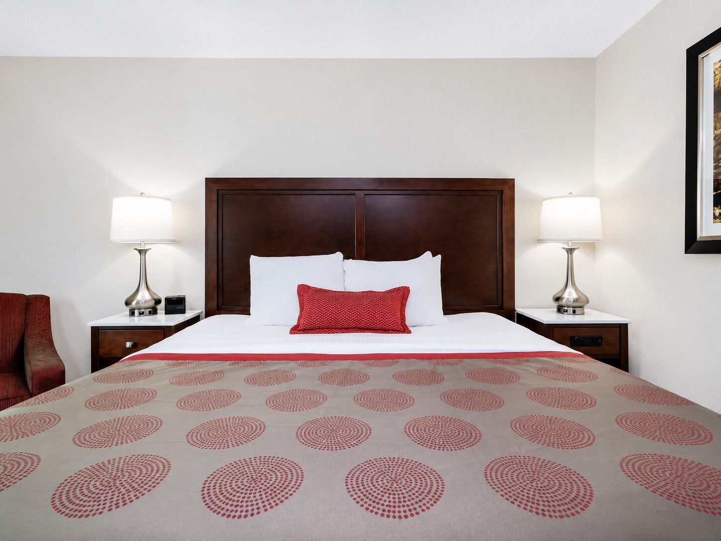 Habitación de hotel con una cama grande, dos mesitas de noche con lámparas, un cojín rojo y una colcha estampada.
