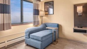 A room with a large window, a blue sofa, a side table, a lamp, a wall-mounted shelf, and a framed picture. - Ramada by Wyndham Minneapolis Golden Valley (Golden Valley)