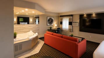 Una habitación de hotel moderna con un sofá grande, una zona de estar separada, un televisor plano grande, una bañera y una mesita con una planta.
