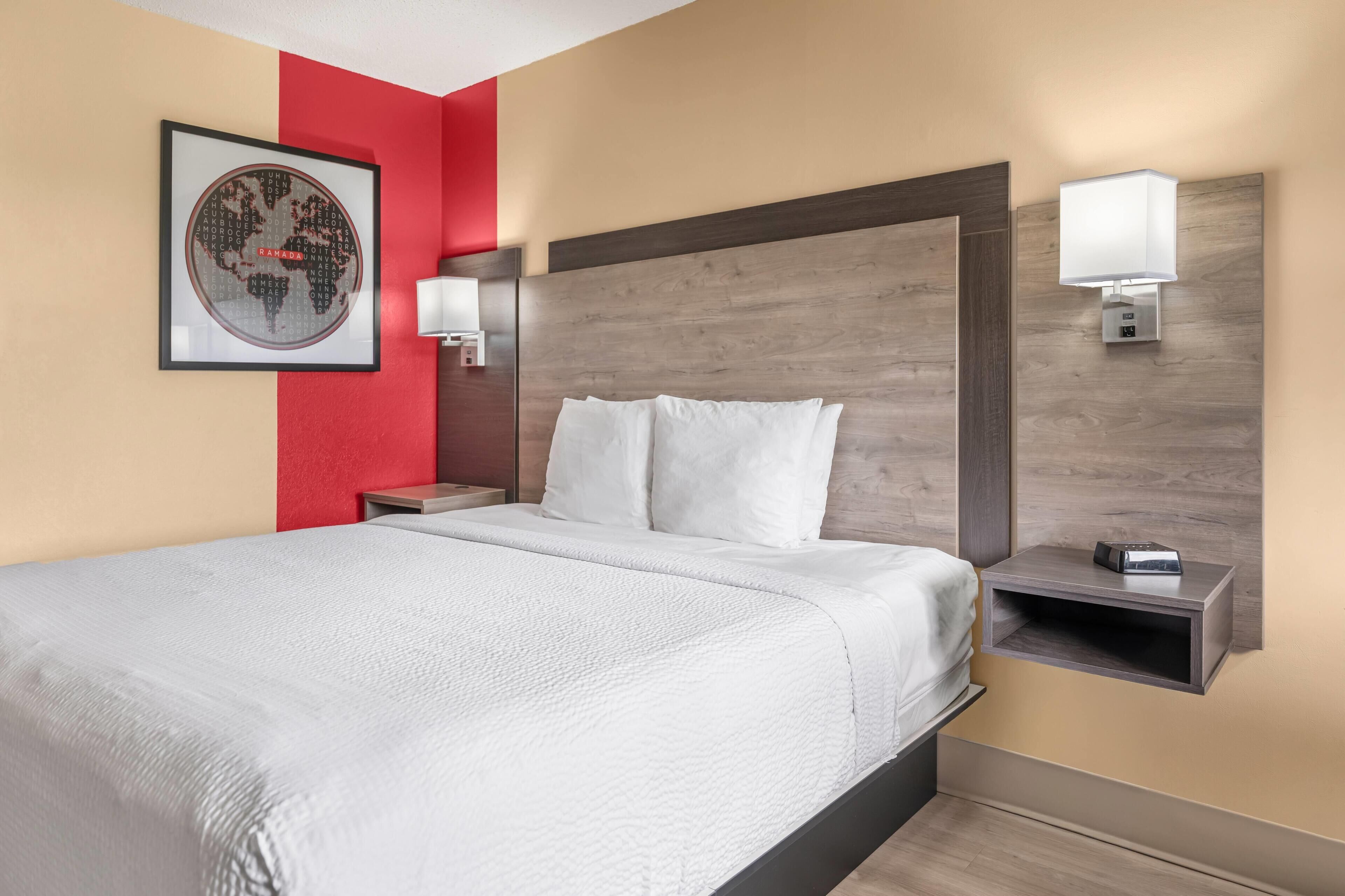 A hotel room with a large bed, a nightstand with a lamp, a wall-mounted shelf, and a framed world map.
