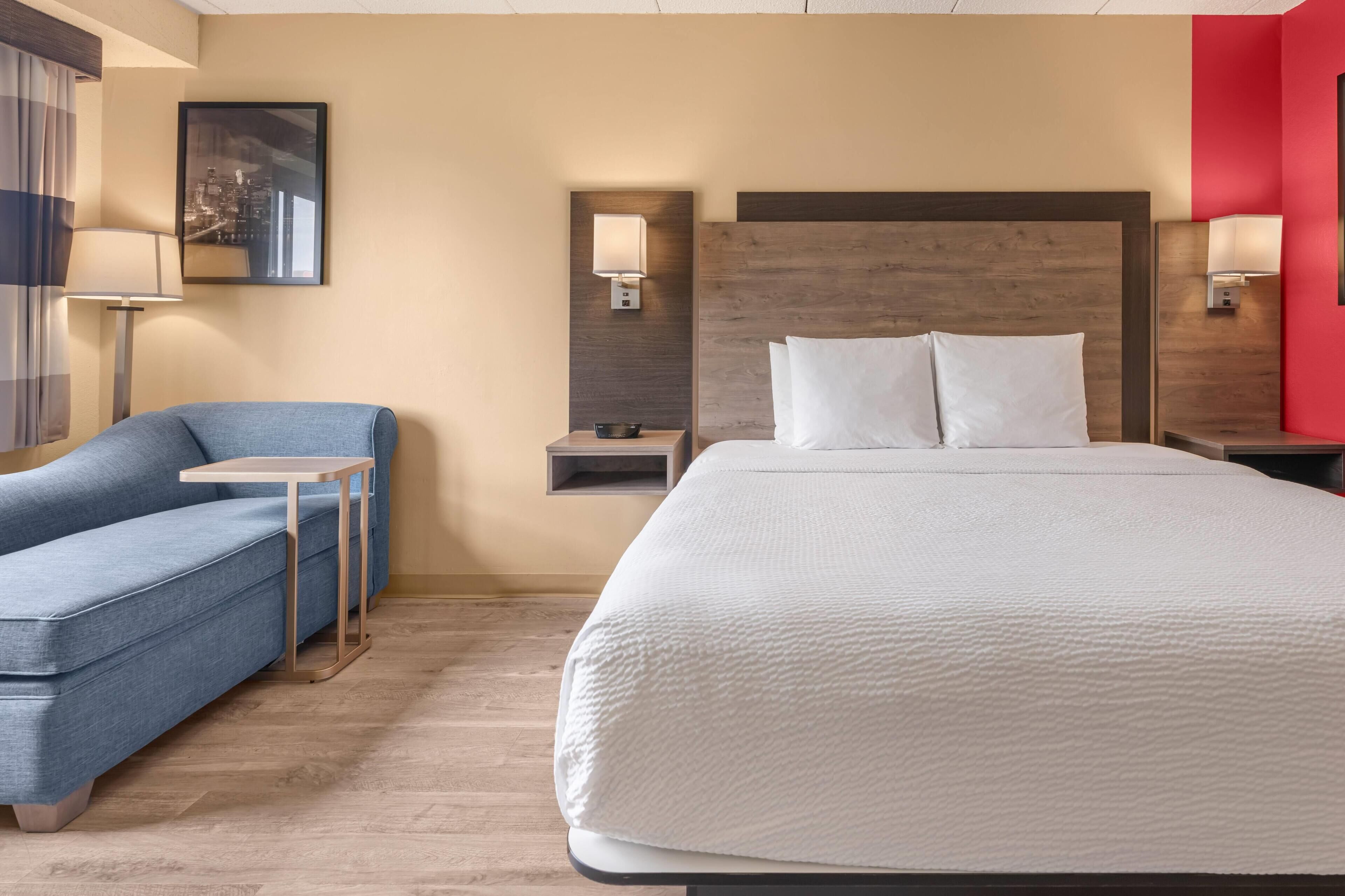 Una habitación de hotel con una cama, un sofá azul, una mesita pequeña y una mesita de noche con lámpara.