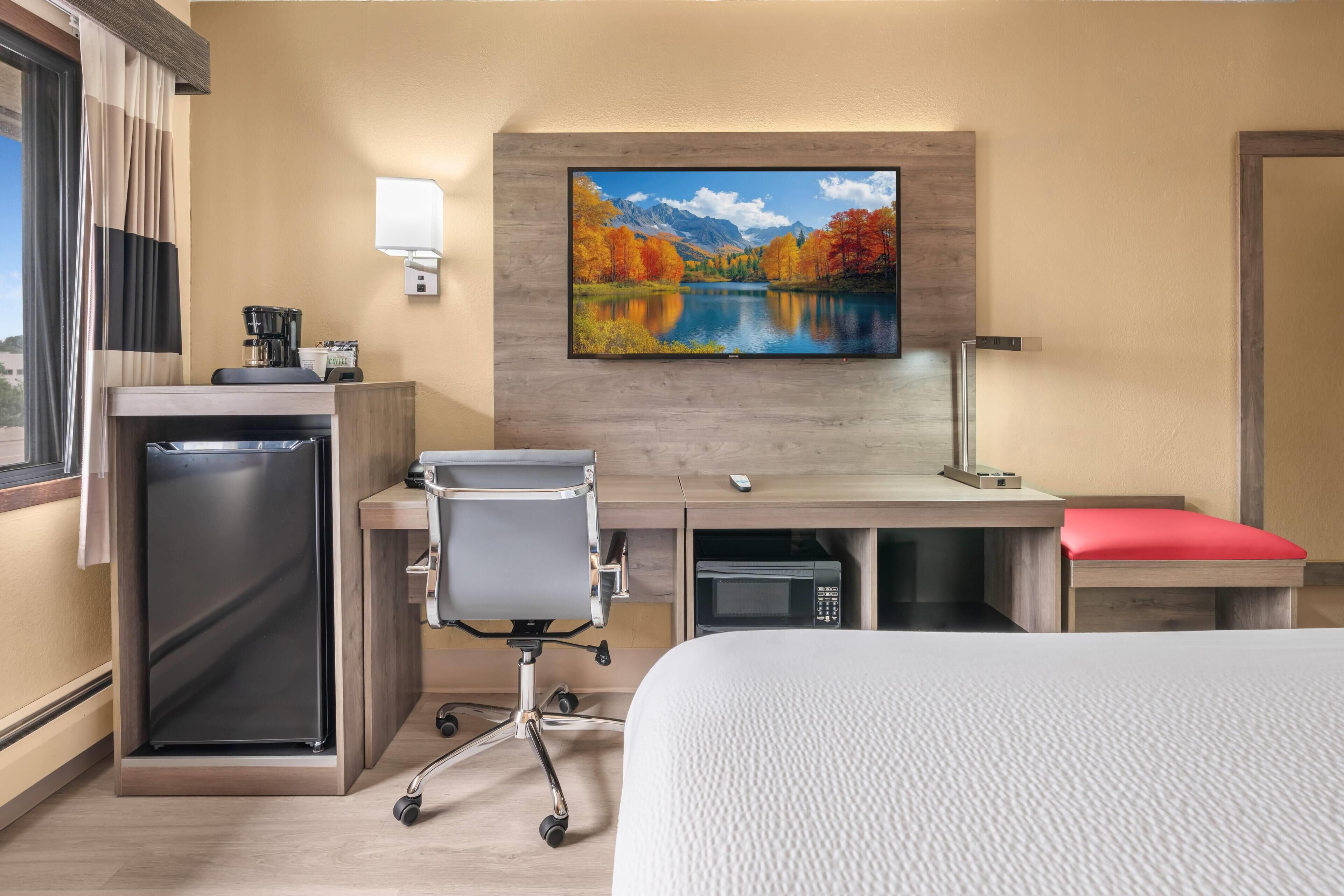 Una habitación de hotel con cama, escritorio, silla, microondas y televisor montado en la pared.