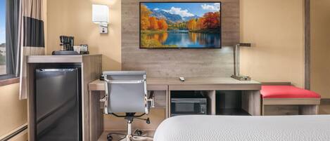 A hotel room with a bed, desk, chair, microwave, and a TV mounted on the wall.