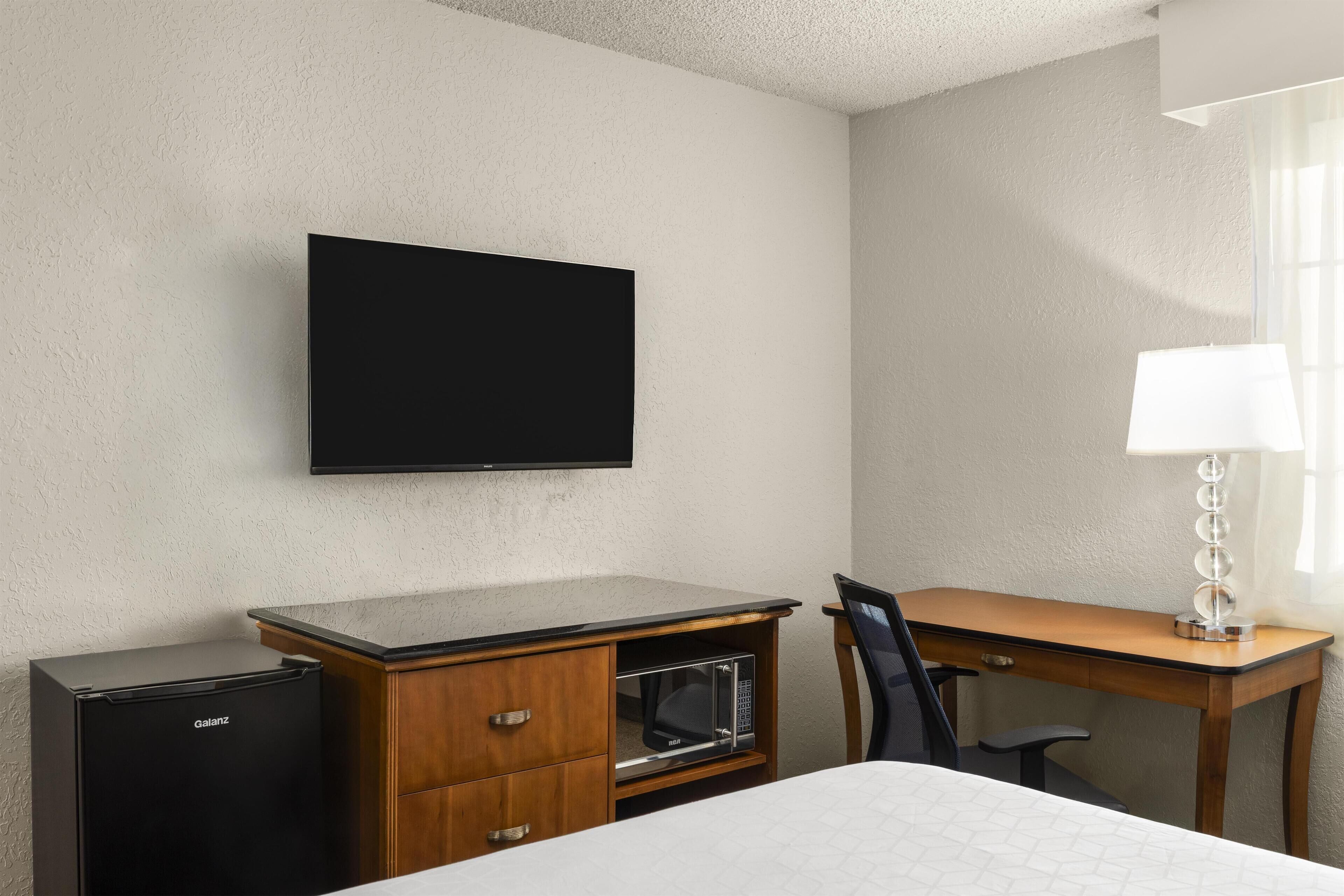 Una habitación de hotel con televisión de pantalla plana montada en la pared, un escritorio de madera con lámpara, microondas y una cama.