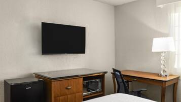 Una habitación de hotel con televisión de pantalla plana montada en la pared, un escritorio de madera con lámpara, microondas y una cama.