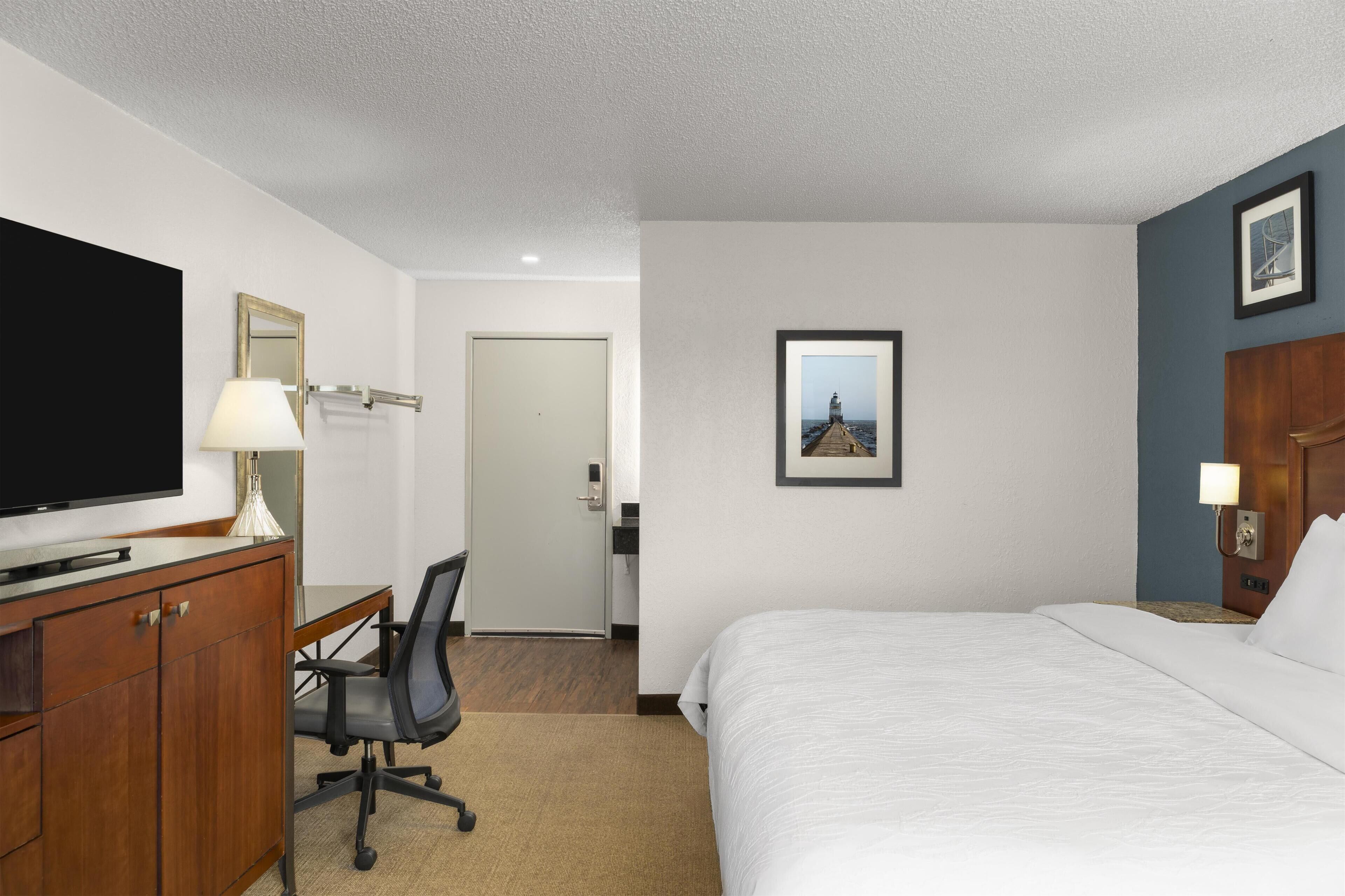 Una habitación de hotel con cama, escritorio, silla, televisor y puerta que da a un baño.