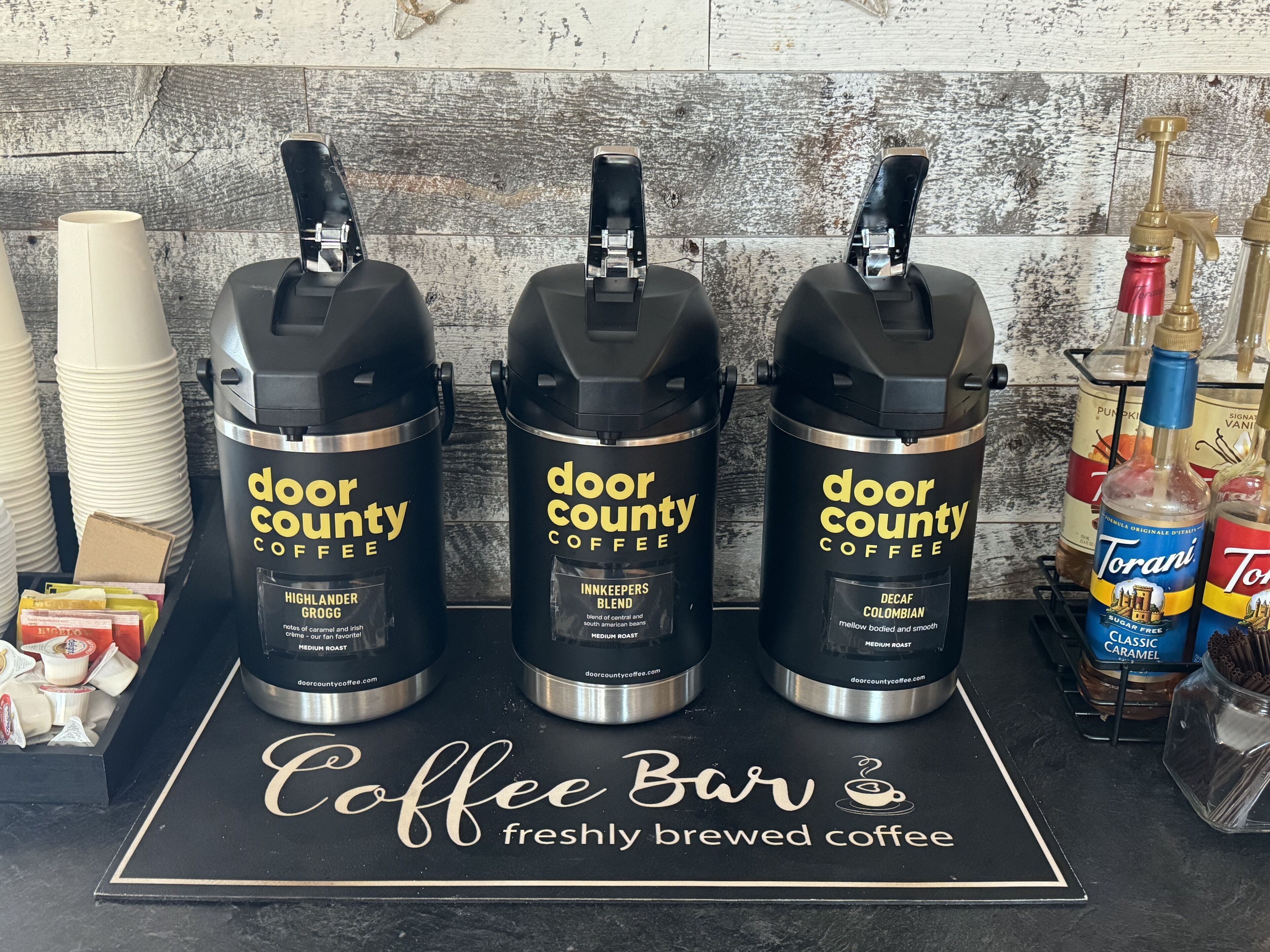 Three coffee dispensers labeled 'door county coffee' with different flavors, a coffee bar sign, and various coffee-related items on a counter.