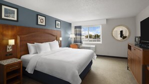 A hotel room with a large bed, a wooden headboard, a nightstand with a lamp, a desk with a TV, and a window with a view of the outdoors. - Harbor Town, BW Signature Collection (Manitowoc)