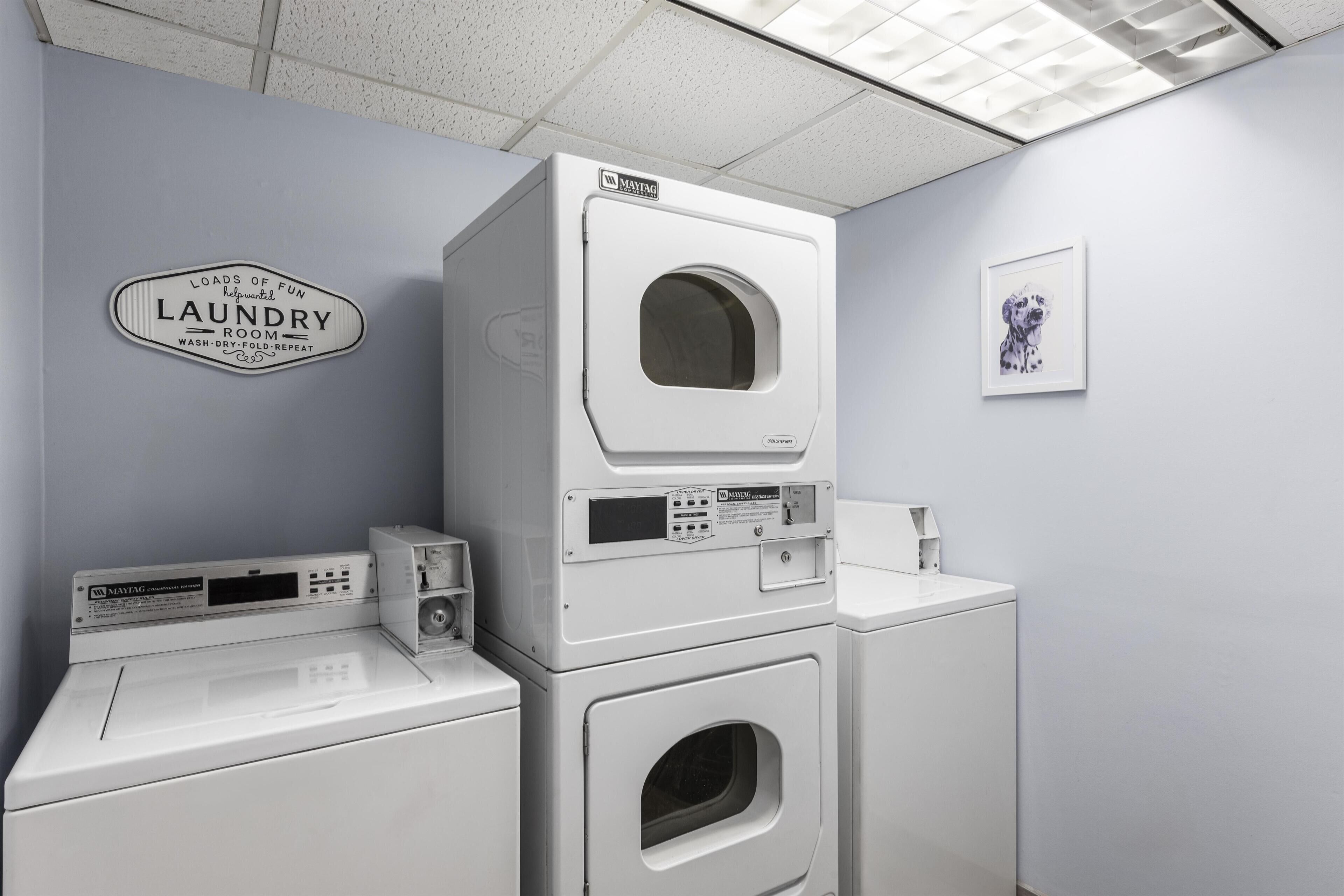 A laundry room with stacked washers and dryers, a sign indicating laundry services, and a framed picture on the wall.