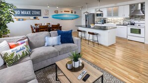 A modern living room with a grey sofa, a coffee table, and a dining area with a wooden table and chairs. A kitchen with white cabinets and stainless steel appliances is visible in the background. - Harbor Town, BW Signature Collection (Manitowoc)