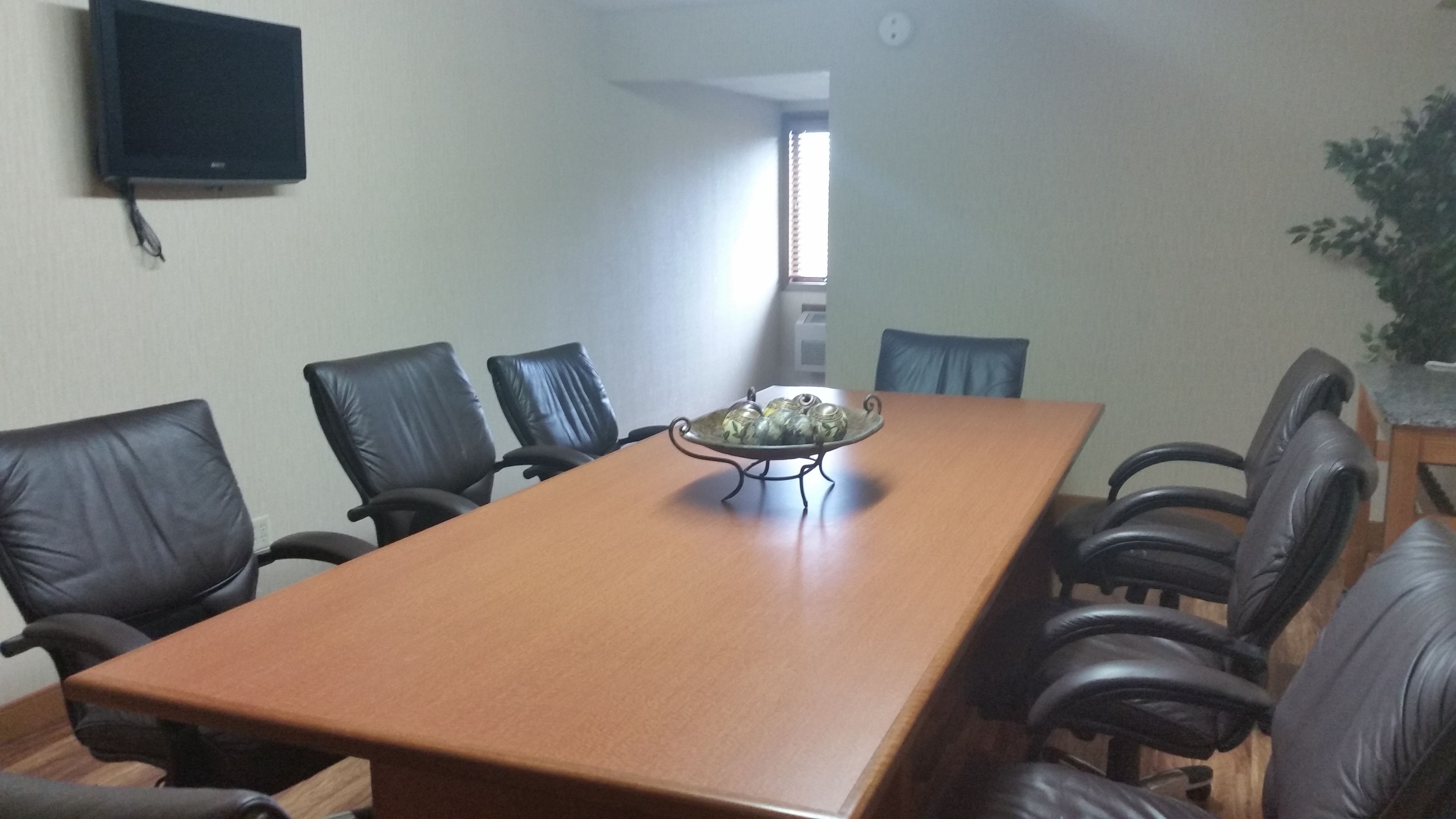 A conference room with a long table, six chairs, a small decorative item on the table, a television on the wall, and a window with blinds.