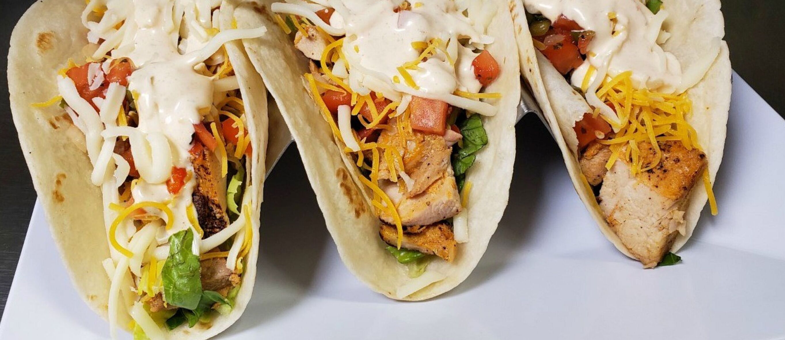 Three tacos with grilled chicken, lettuce, tomatoes, and shredded cheese, topped with a creamy sauce.