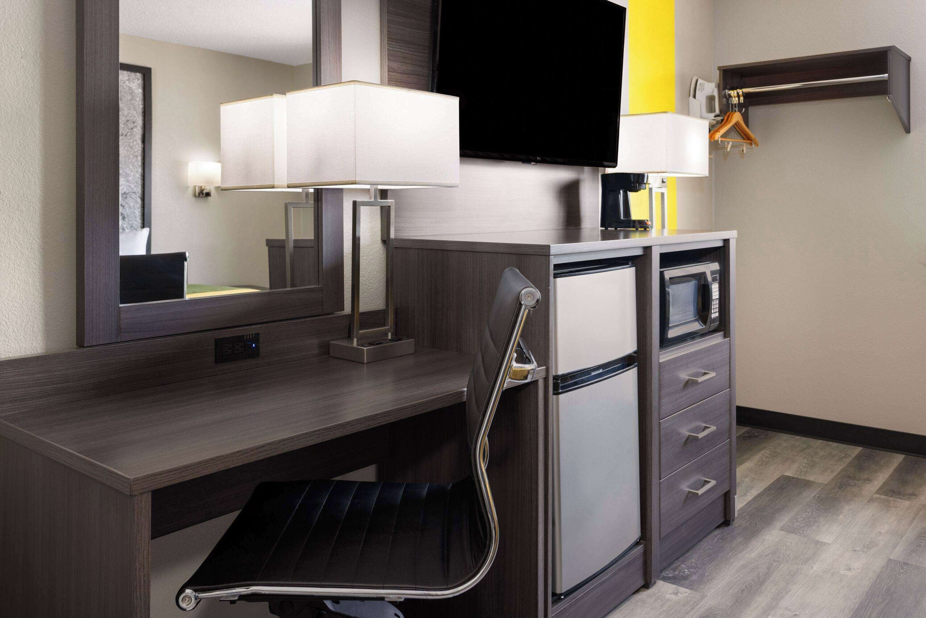 A hotel room with a dark wood desk, a mirror, a flat-screen TV, a small refrigerator, a microwave, a sink with a towel, and a shower area.