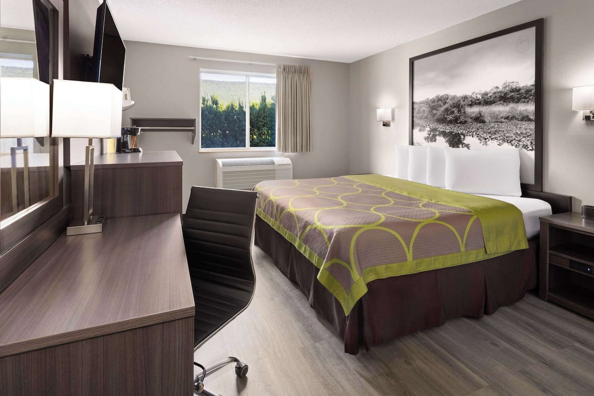 Ein Hotelzimmer mit Bett, Schreibtisch, Stuhl, einem Fenster mit Vorhängen, einem an der Wand befestigten Fernseher und einem großen gerahmten Bild.
