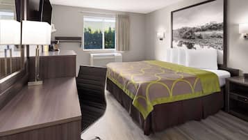 A hotel room with a bed, a desk, a chair, a window with curtains, a wall-mounted TV, and a large framed picture.