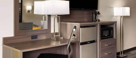 A hotel room with a desk, a microwave, a coffee maker, a TV, and a chair.