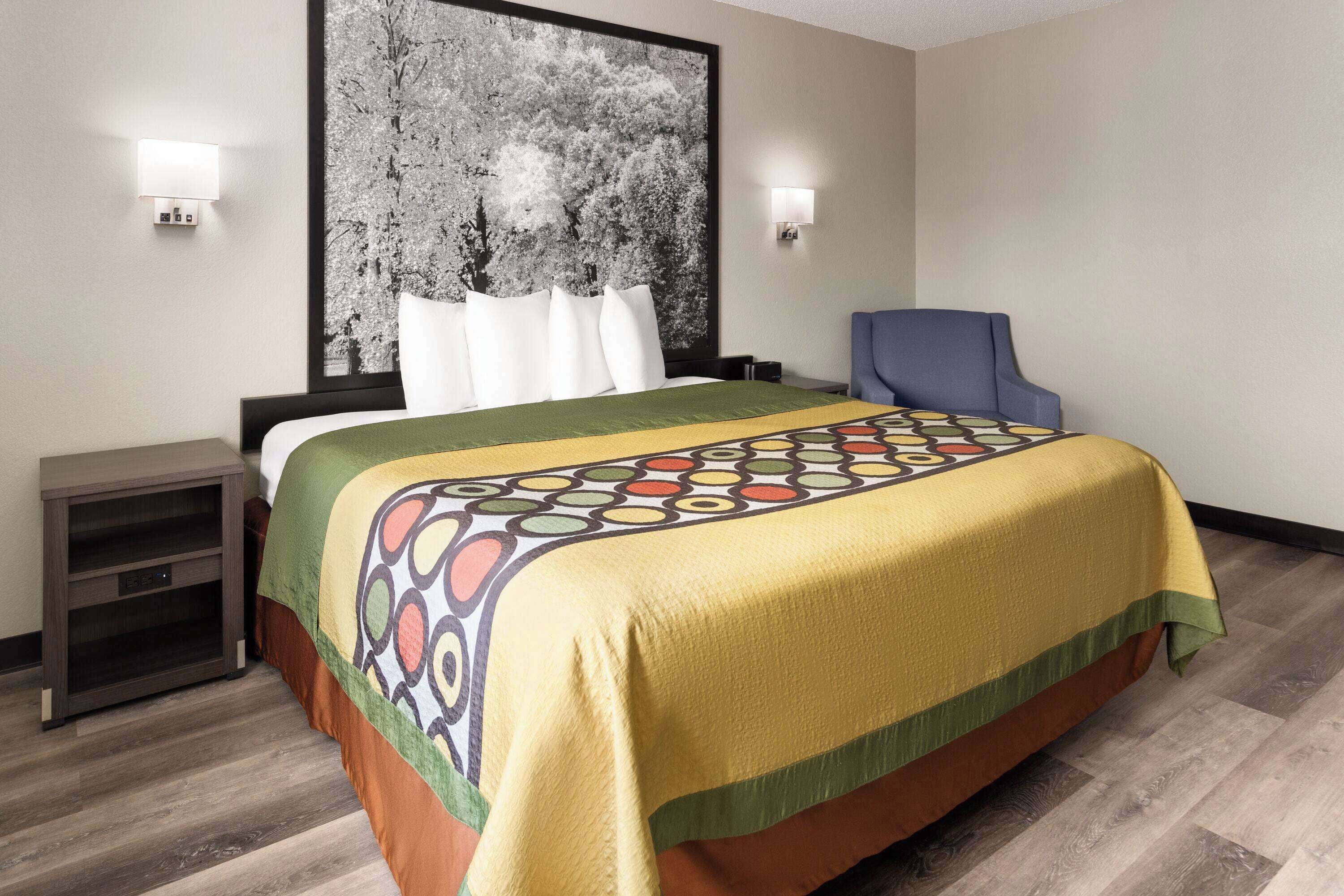 A hotel room with a large bed, a chair, a nightstand, and a wall-mounted lamp.