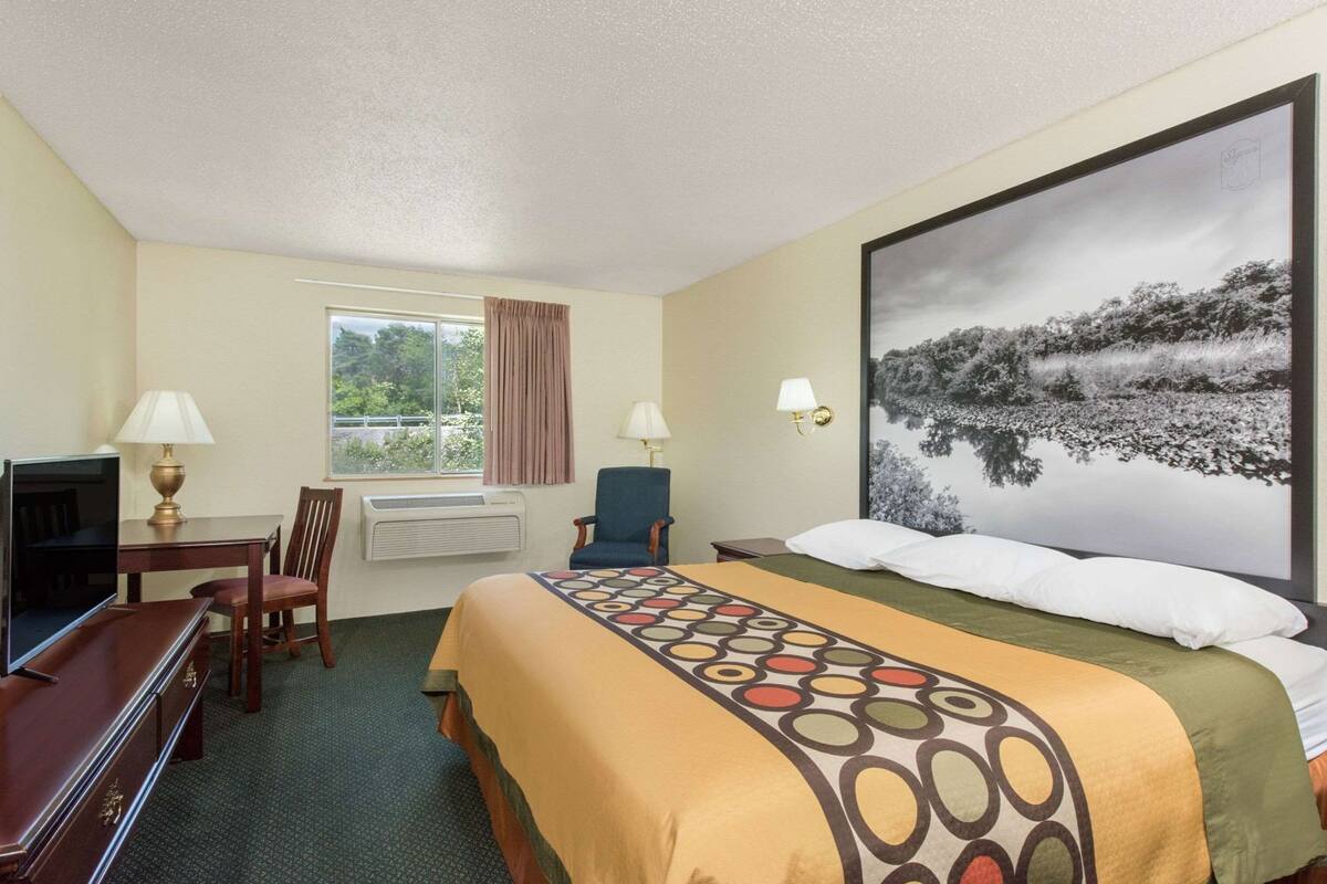 Ein Hotelzimmer mit einem Bett, einem Schreibtisch mit Fernseher, einem Sessel, einem Fenster mit Vorhängen und einem großen Gemälde an der Wand.